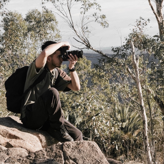 A photographer seated on a rock, holding a camera to his eye among trees.