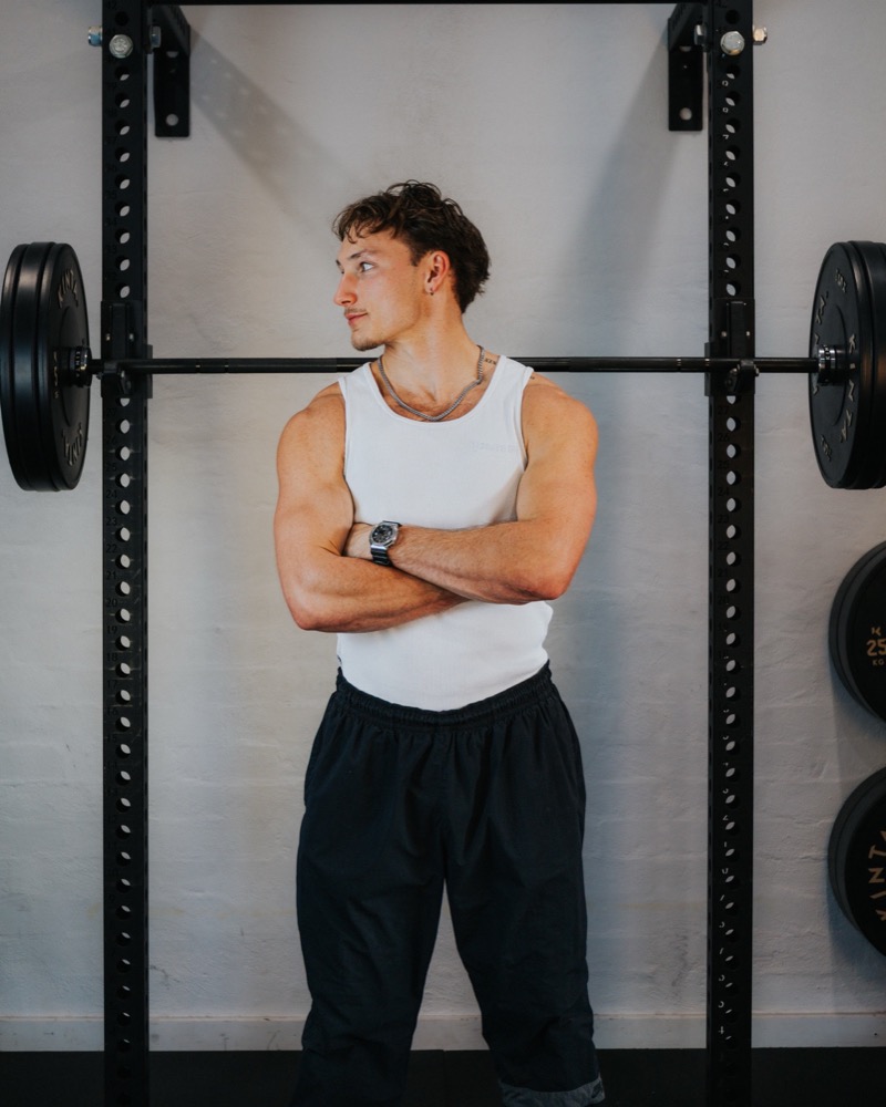 Jackson Pitsakis standing with his arms folded in a white sleeveless top beside a gym squat rack.