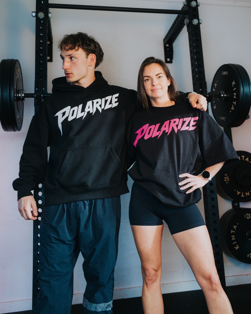 Two people wearing a black Polarize hoodie and a black T-shirt with pink lettering beside a squat rack.