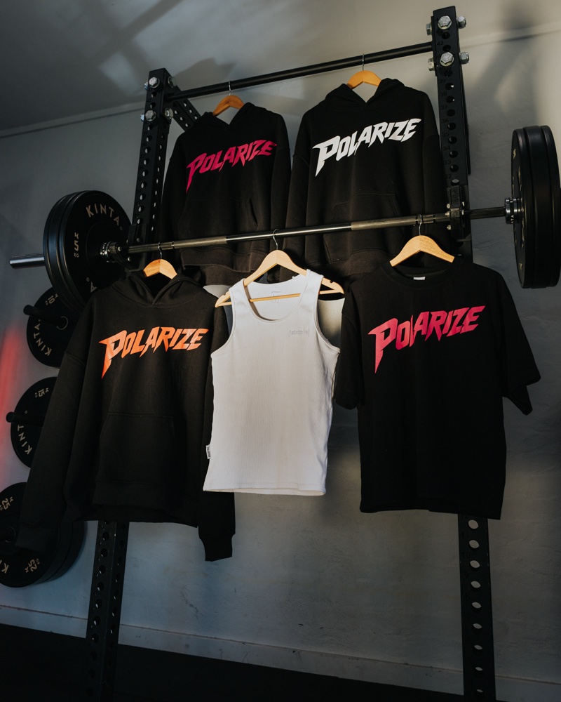 Polarize hoodies and T-shirts hanging from a gym squat rack, with pink, white and orange lettering.