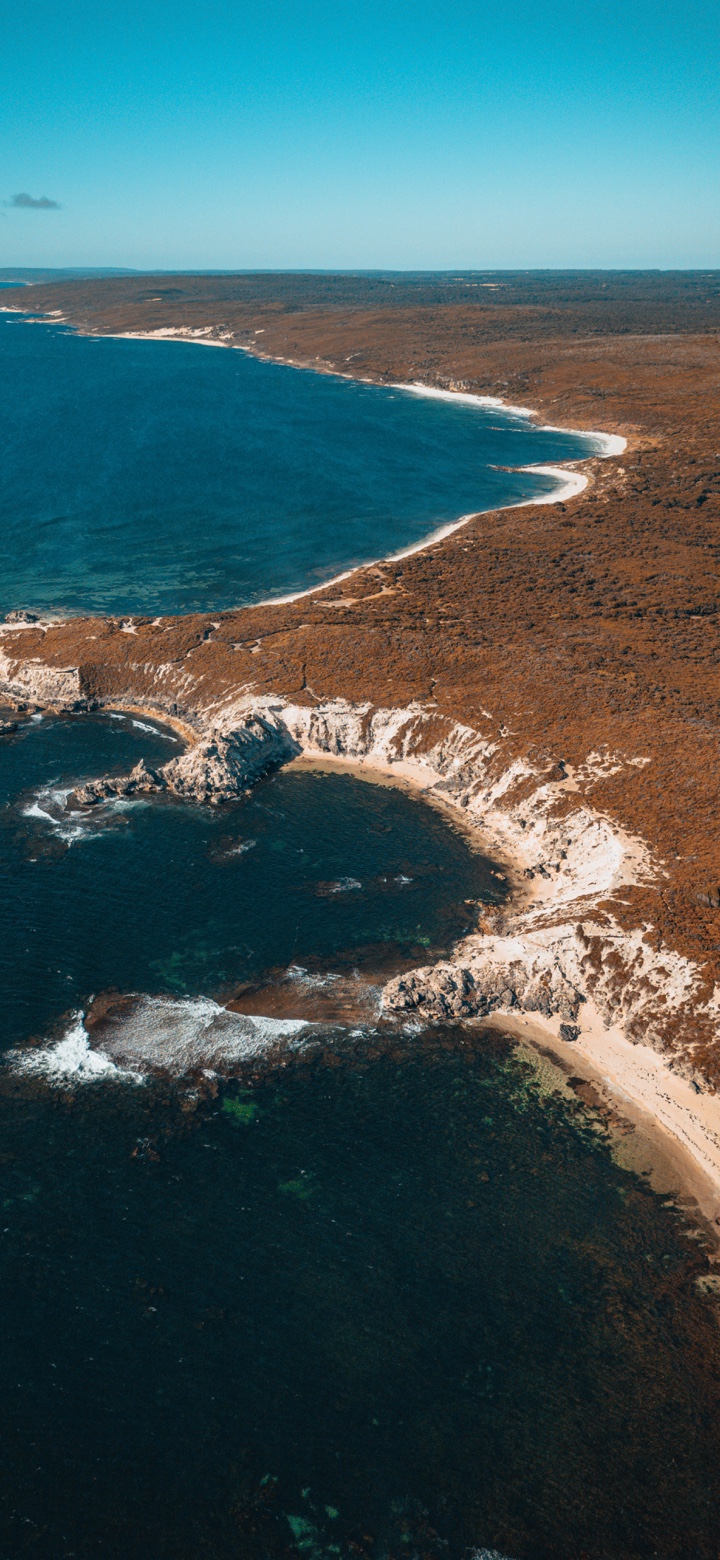 An aerial view of pale cliffs curling around deep blue coves beneath ochre coastal scrubland.