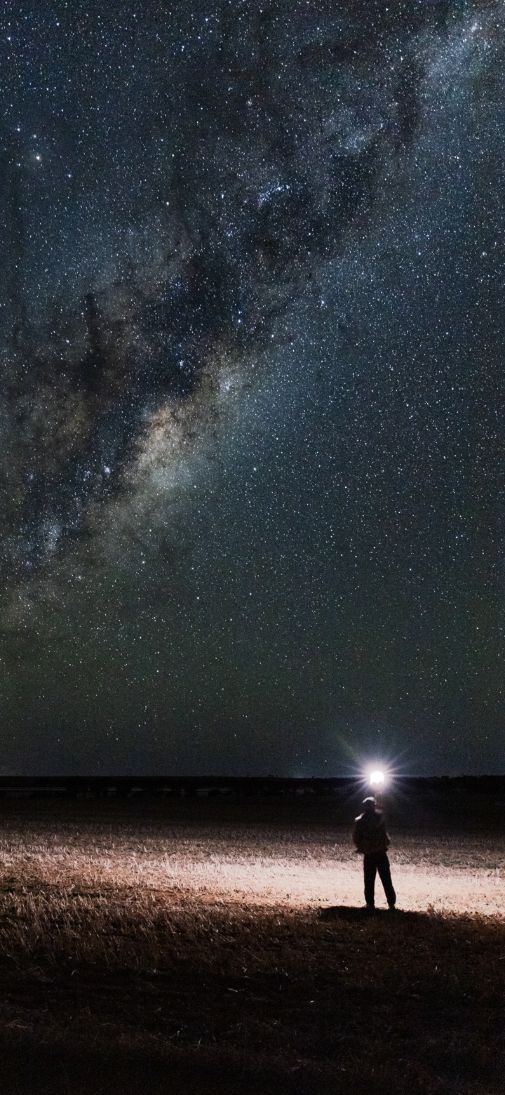 The Milky Way above a person standing in a dark field beneath a bright light.